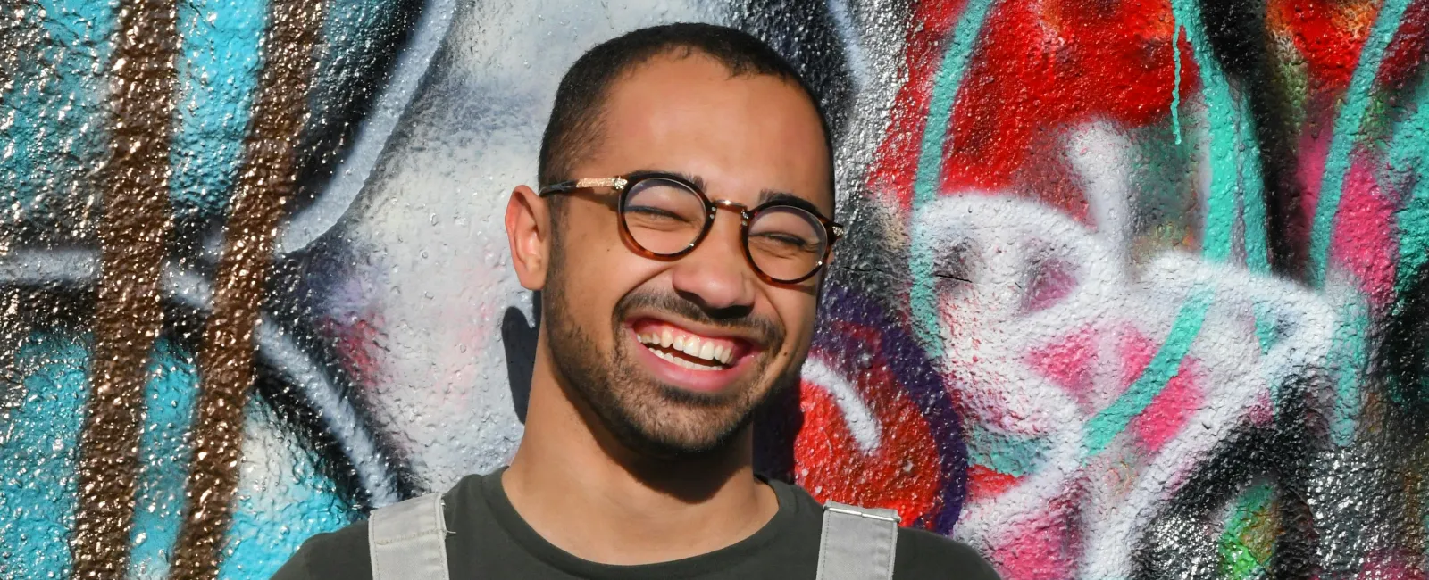 Smiling man with glasses wearing grey overalls and holding a black cap stands against colorful graffiti wall.