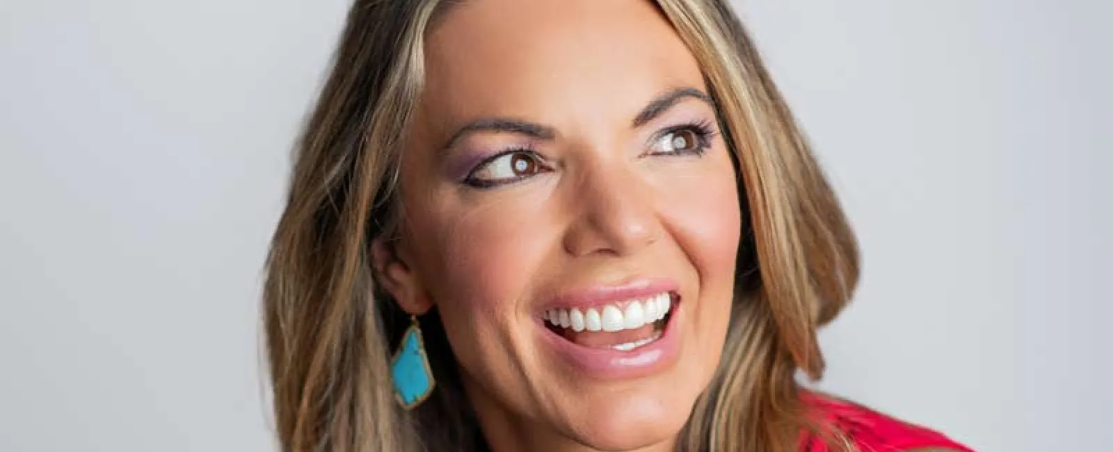 Smiling woman with long blonde hair wearing turquoise earrings and a red sleeveless top, showcasing a bright smile.