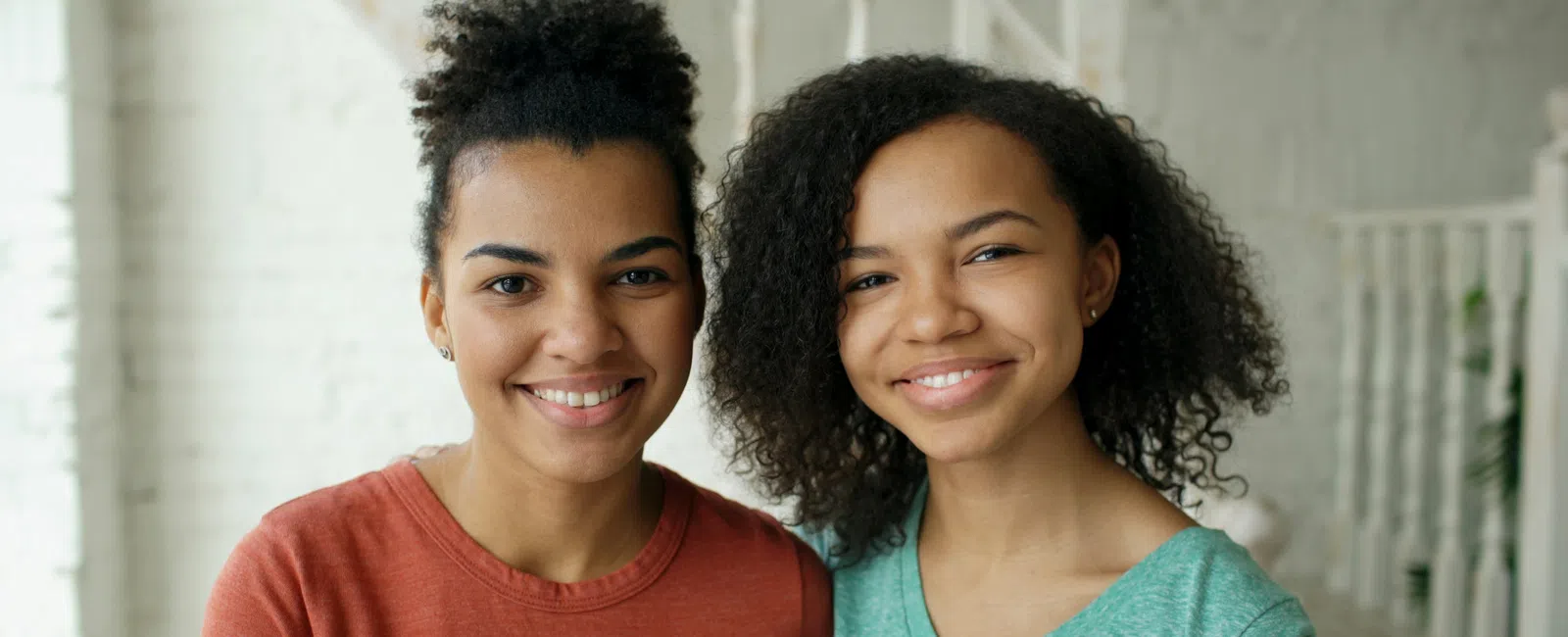 Two smiling young women with curly hair standing close together in casual clothes indoors