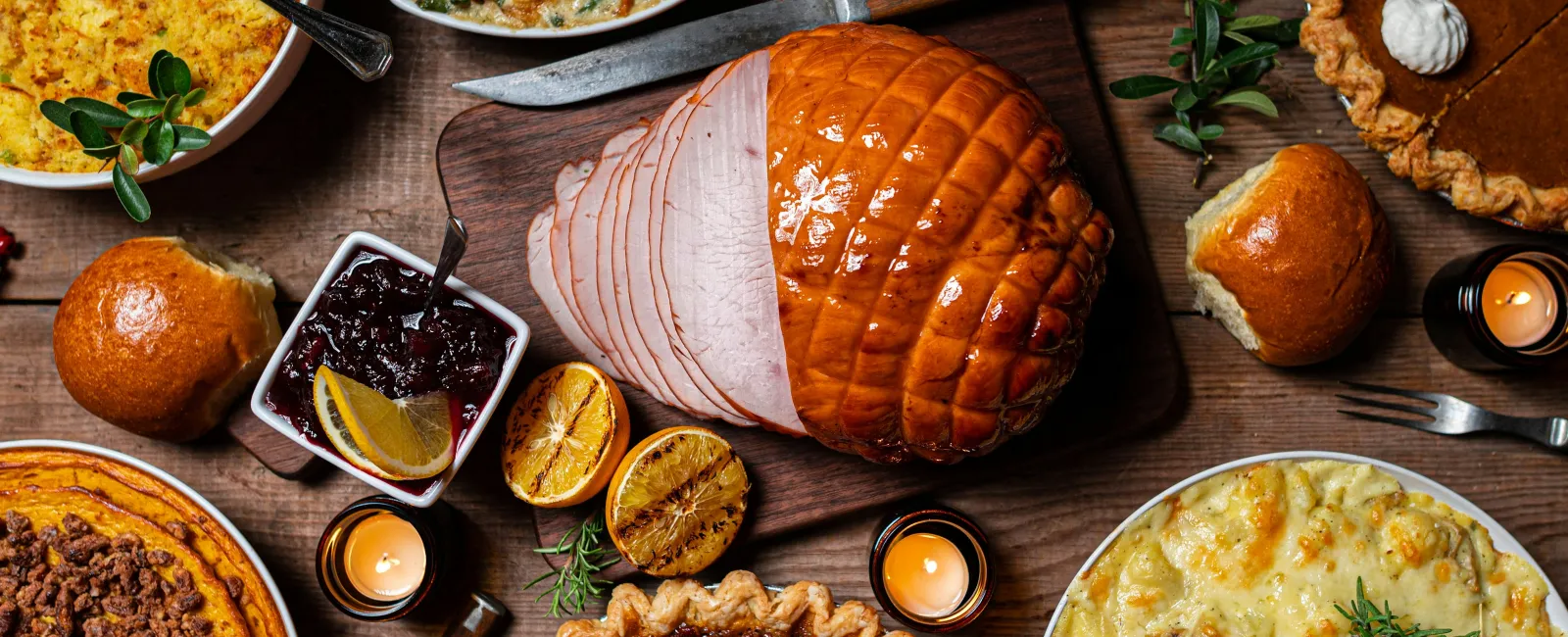 Traditional Thanksgiving feast with glazed ham, pecan pie, pumpkin pie, casseroles, dinner rolls, and gravy on wooden table.