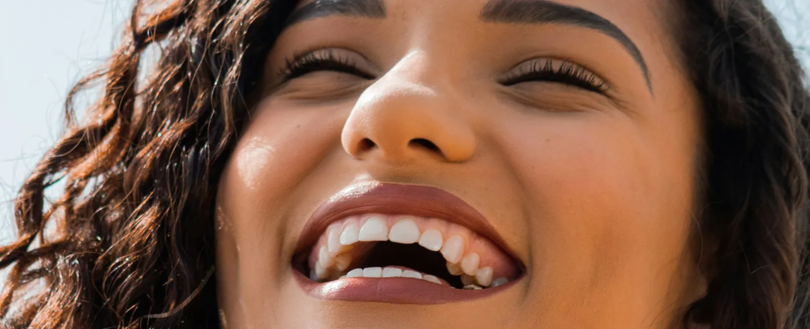 Close-up of a young woman with curly hair smiling brightly against a clear blue sky background.