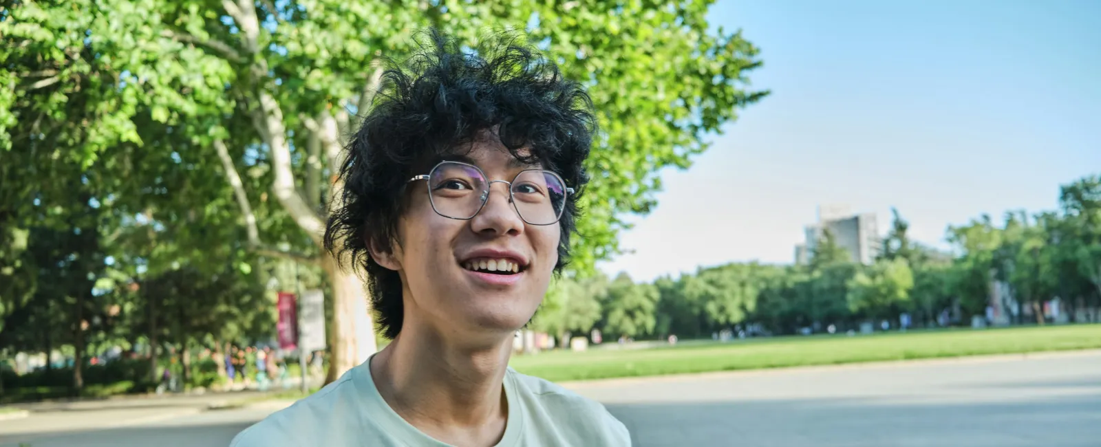 Young man with glasses smiling outdoors in a park setting on a sunny day with trees and green grass.