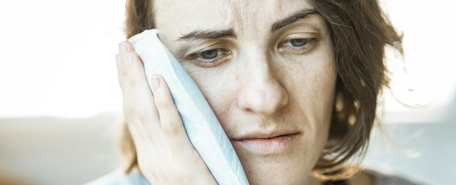 Woman with a pained expression holding an ice pack to her cheek outdoors in daylight