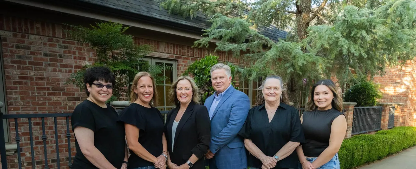 Six diverse professionals standing outdoors in front of a brick building with greenery, smiling at the camera.
