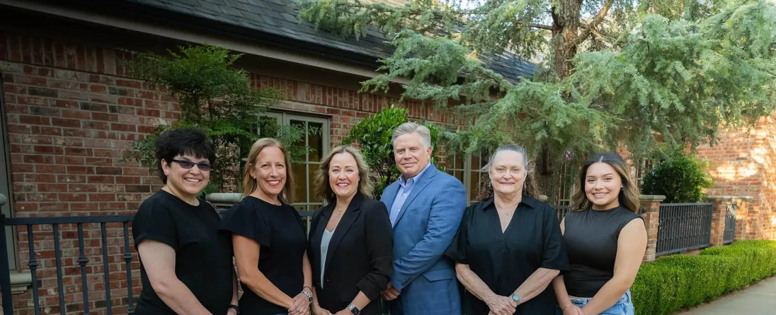 Six diverse professionals standing outdoors in front of a brick building with greenery, smiling at the camera.