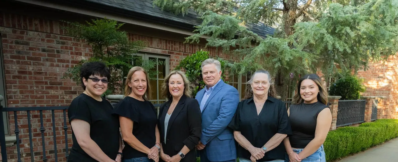 Six diverse professionals standing outdoors in front of a brick building with greenery, smiling at the camera.