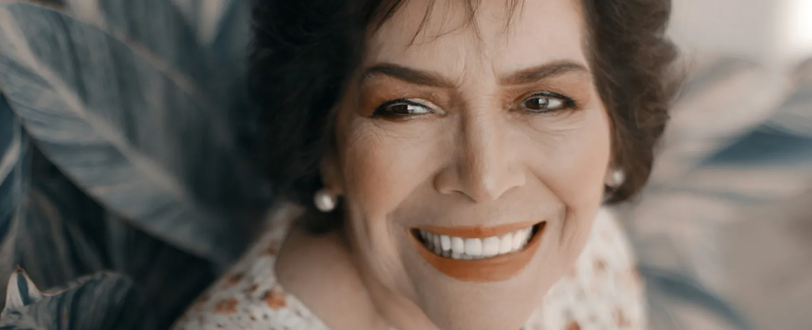 Smiling mature woman with short hair wearing a floral blouse against leafy background outdoors.