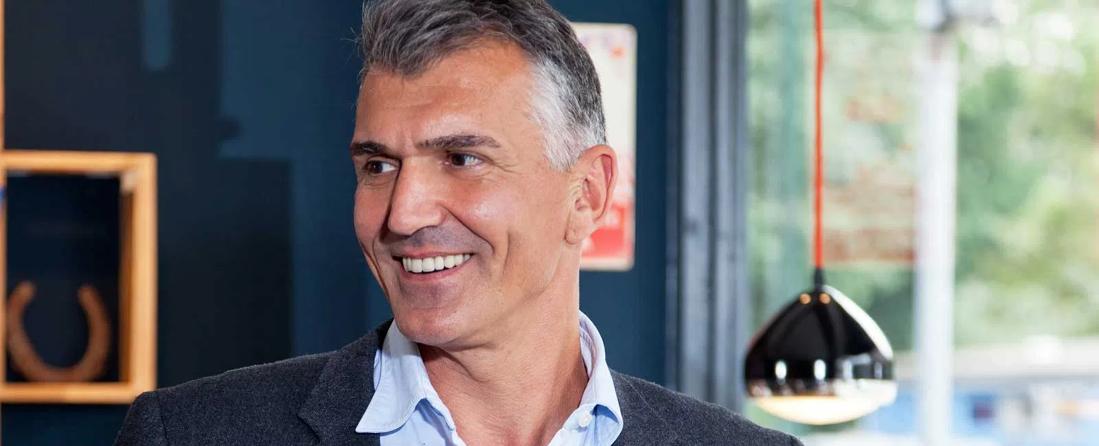 Smiling mature man in a gray blazer and blue shirt standing indoors near a window with modern decor.
