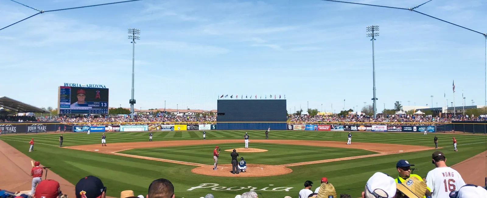 Baseball game in progress at Peoria stadium with fans watching under clear blue sky and bright sunlight