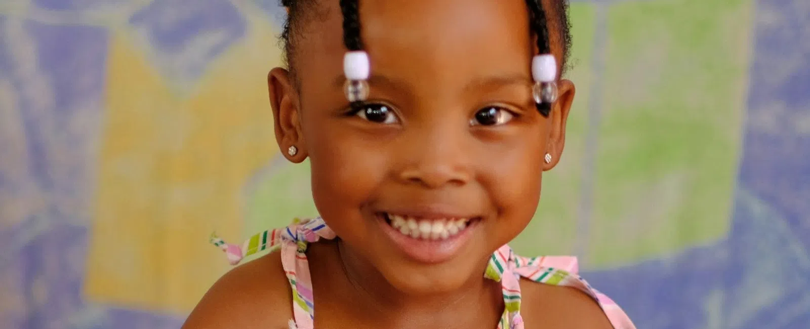 Smiling little girl in a colorful striped dress sitting cross-legged against a multicolor abstract background.