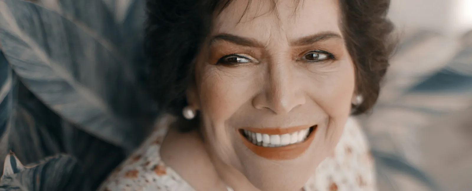 Smiling mature woman with short dark hair wearing floral blouse and pearl earrings in soft natural light.
