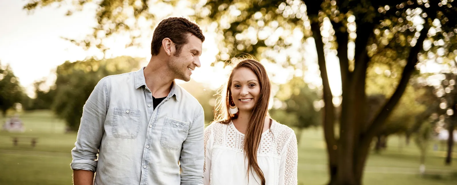 Young couple holding hands and smiling in a sunlit park with trees and grass in the background