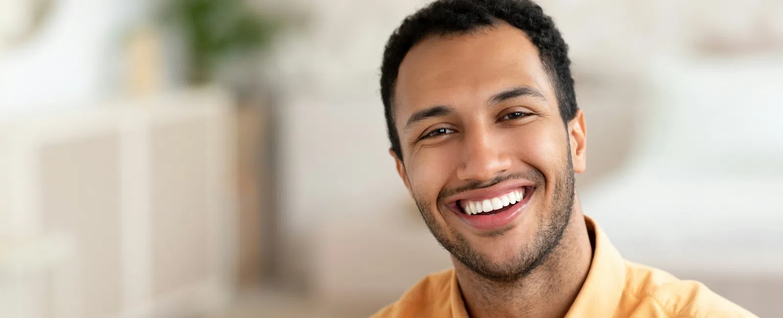 Smiling young man wearing a mustard shirt sitting indoors with a blurred background, looking happy and approachable
