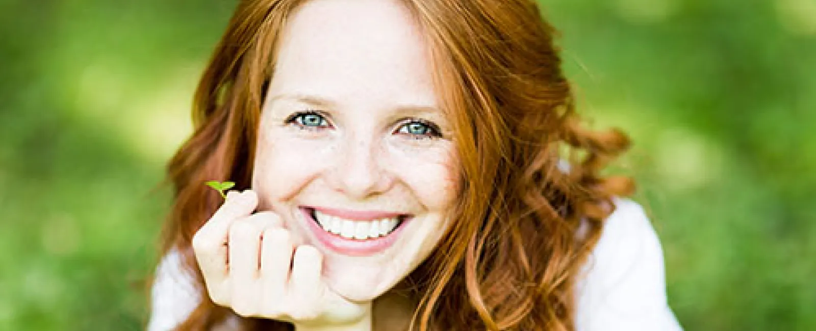 Smiling red-haired woman with curly hair and blue eyes resting chin on hand in a green outdoor setting.