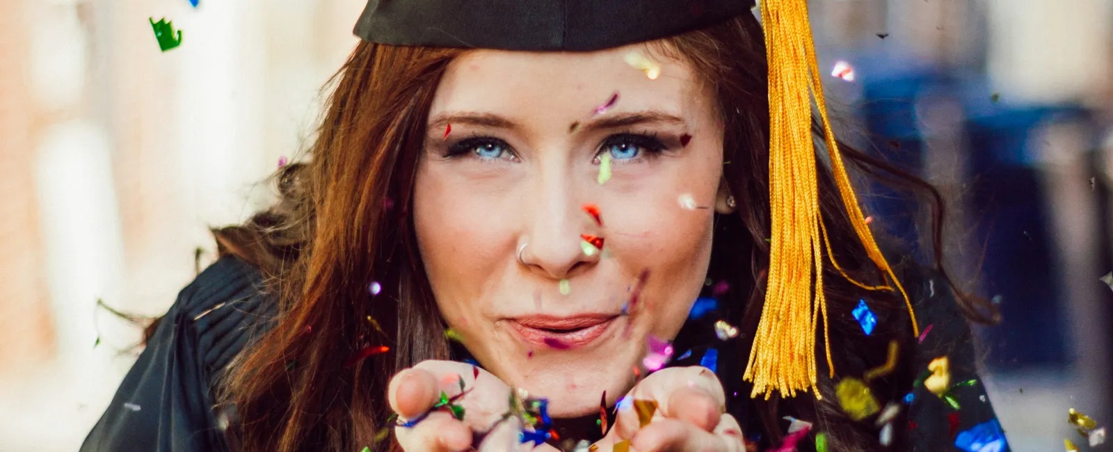 Female graduate wearing cap and gown blows colorful confetti celebrating graduation day outdoors.