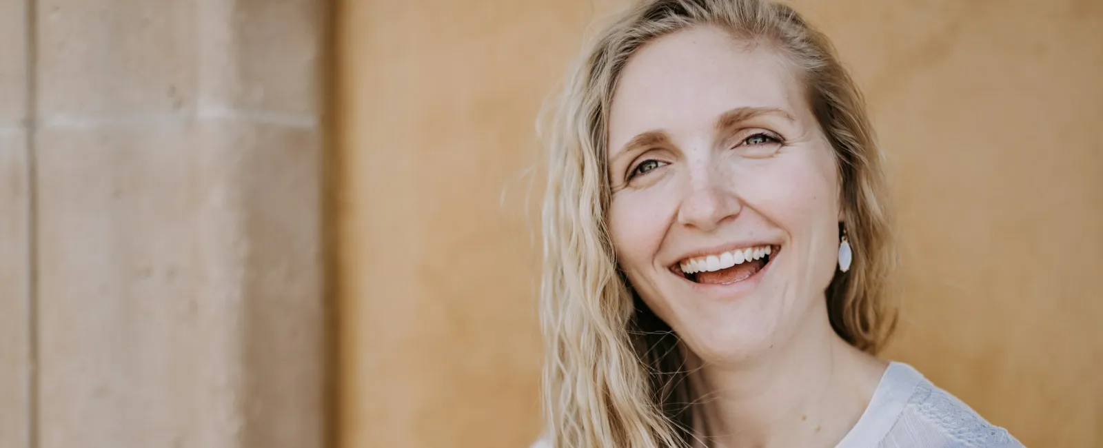 Smiling blonde woman with long wavy hair wearing white lace top against beige textured wall.