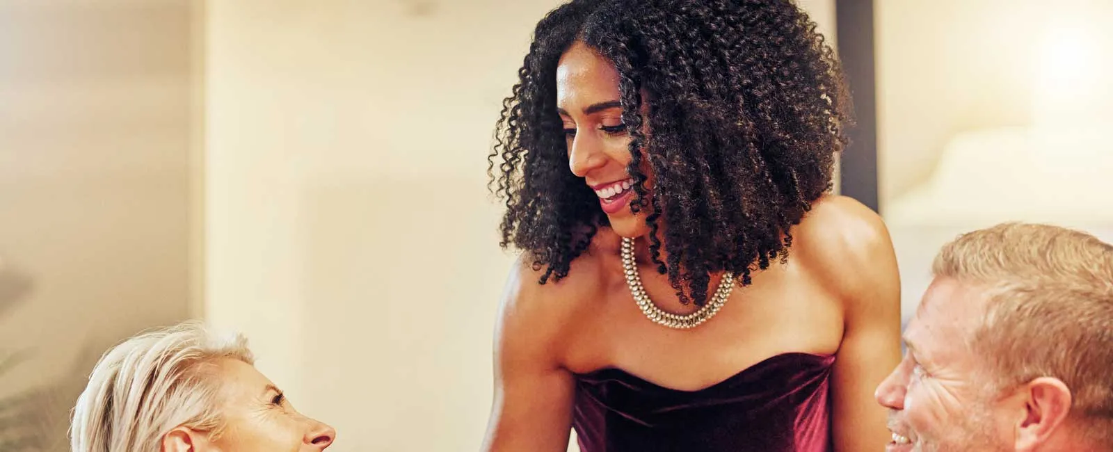Smiling woman in elegant velvet dress chatting with two formally dressed guests at a cozy dinner party.