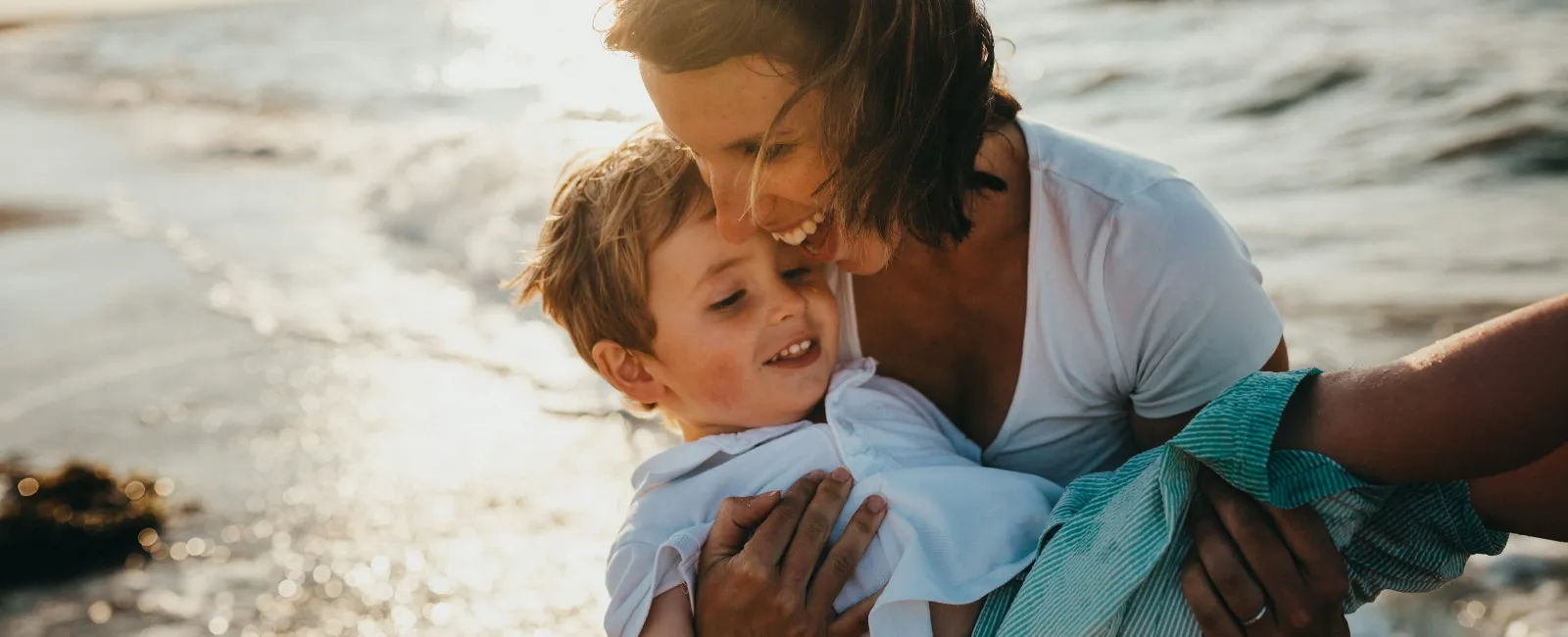 Woman holding and smiling with young boy at the beach during sunset near the ocean shore.