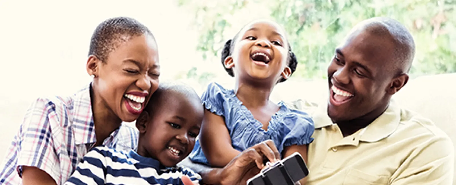 Happy African American family of four laughing and enjoying time together while using a smartphone indoors.