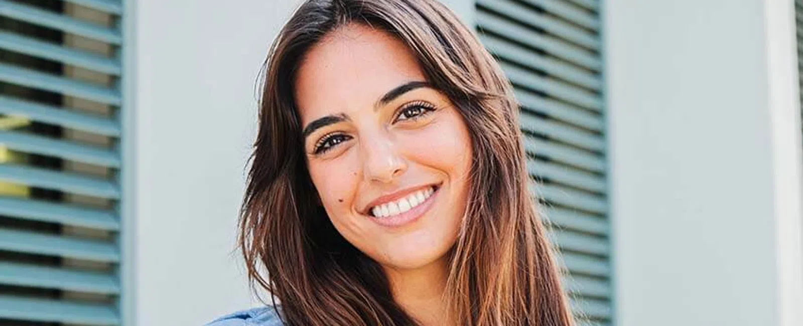 Smiling young woman with long hair wearing a blue shirt, standing in front of green shutters.