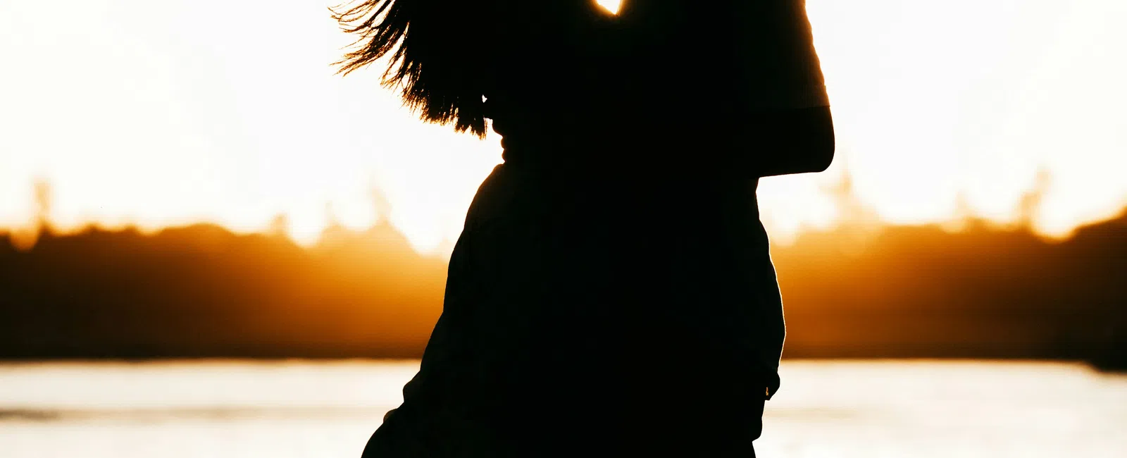 Silhouetted couple embracing at sunset by the water with flowing dress and glowing horizon backdrop