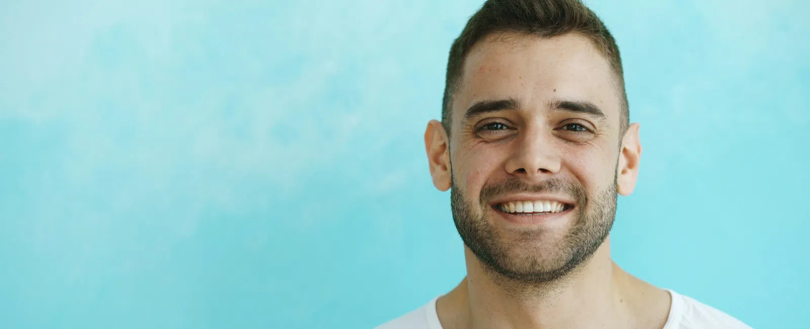 Smiling young man with short hair and beard wearing a white shirt against a light blue background.