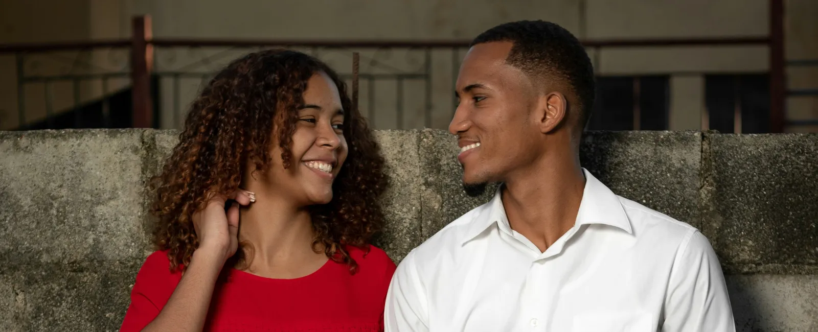 Smiling couple holding hands and leaning against a concrete wall, dressed in black and white and red clothing.