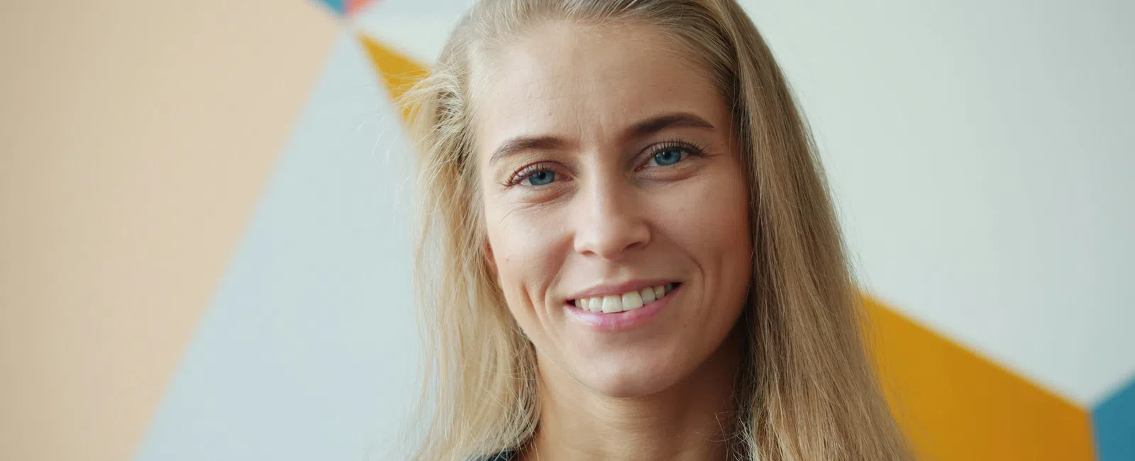Smiling young woman with long blonde hair wearing a denim shirt in front of a colorful geometric wall.