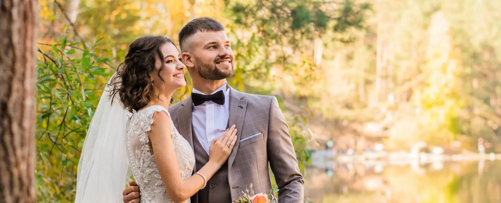 Bride and groom in wedding attire embracing near a serene lake surrounded by autumn trees and soft sunlight.