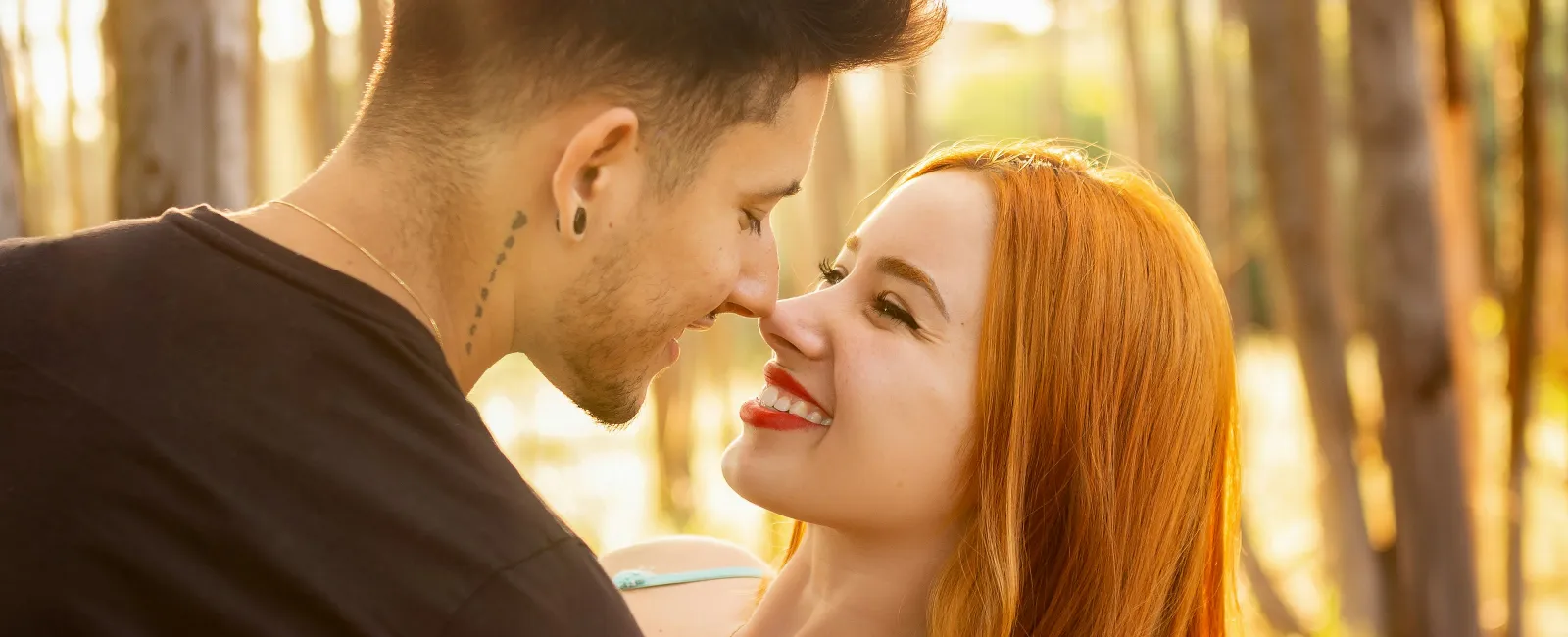 Young couple embracing and smiling at each other in a sunlit forest during golden hour.