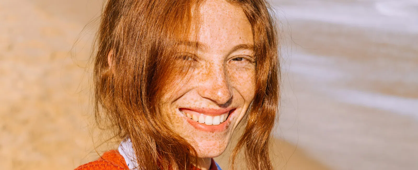 Smiling red-haired woman with freckles wearing an orange sweater on a sunny beach.