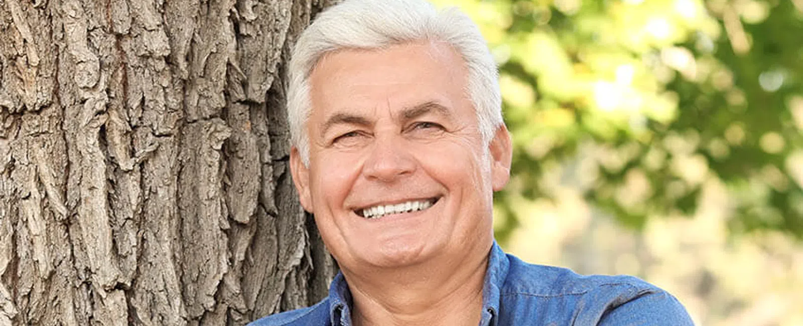 Smiling senior man in a denim shirt leaning against a tree, exuding warmth and confidence in natural light.
