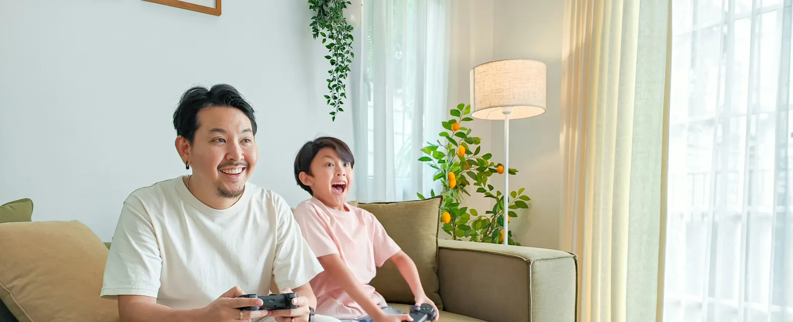 Father and son playing video games together on a sofa in a bright, cozy living room with plants and natural light.