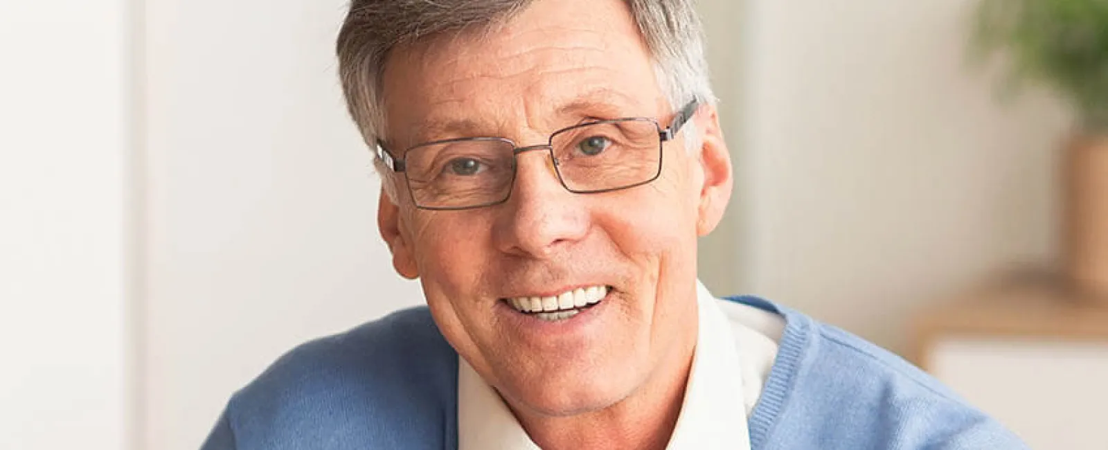 Smiling older man in a blue sweater, glasses, sitting indoors with natural light, exuding warmth and friendliness.