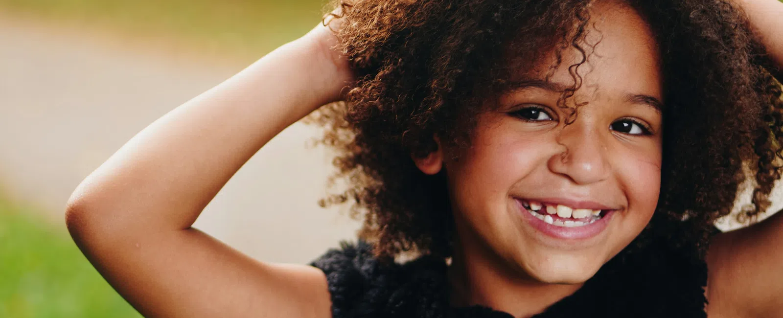 Smiling curly-haired child outdoors with hands in hair wearing a black vest on a blurred green background