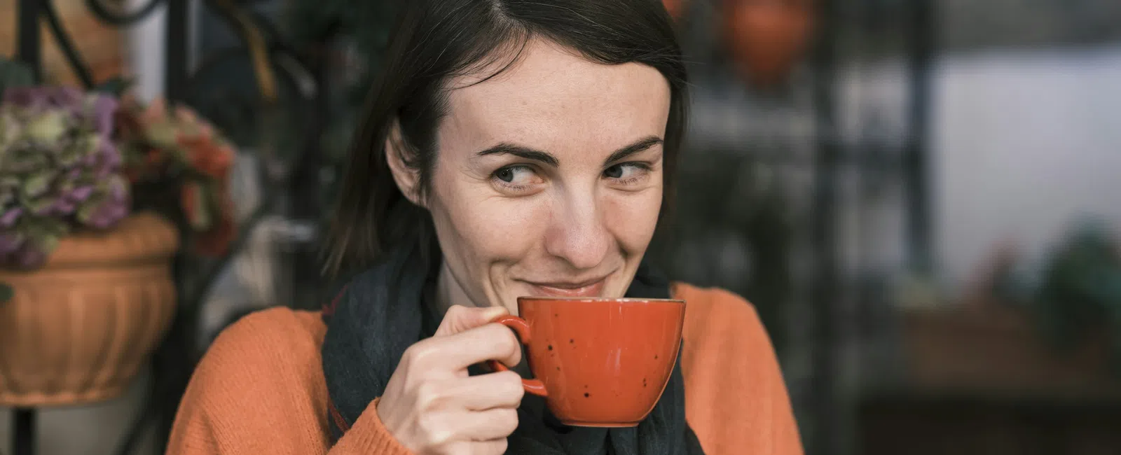 Woman in orange sweater sipping coffee from red cup with a playful smile in cozy cafe setting.