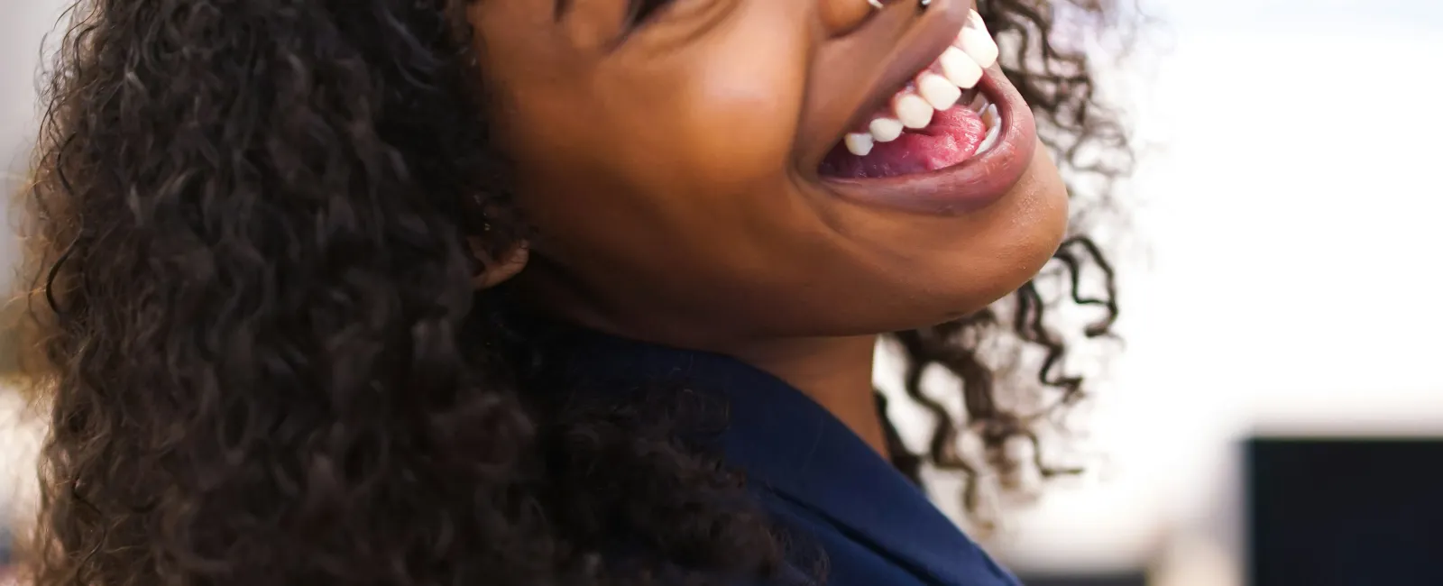 Smiling woman with curly hair and nose ring wearing a sleeveless navy top in a bright room