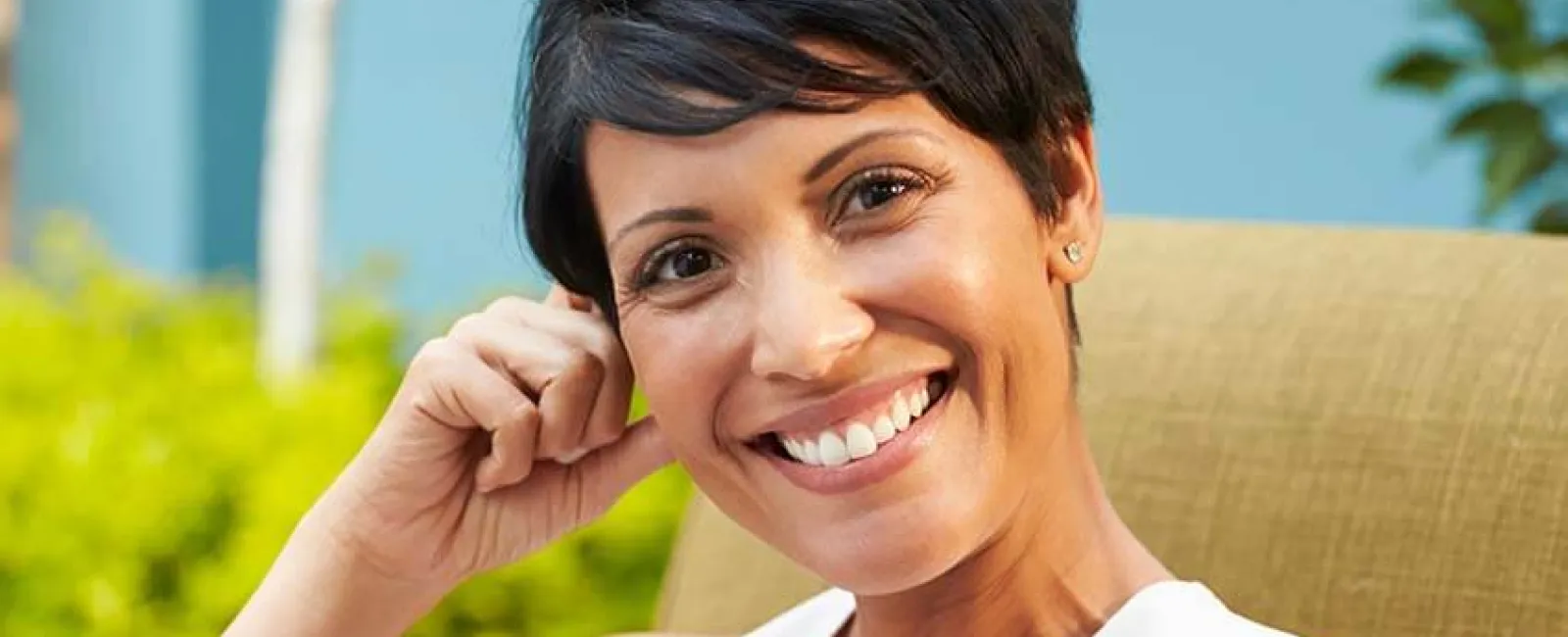 Smiling woman with short hair sitting outdoors, enjoying a sunny garden setting.