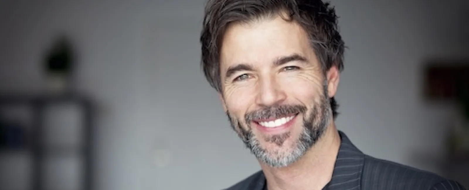 Smiling confident man with salt and pepper beard wearing a dark pinstripe blazer indoors