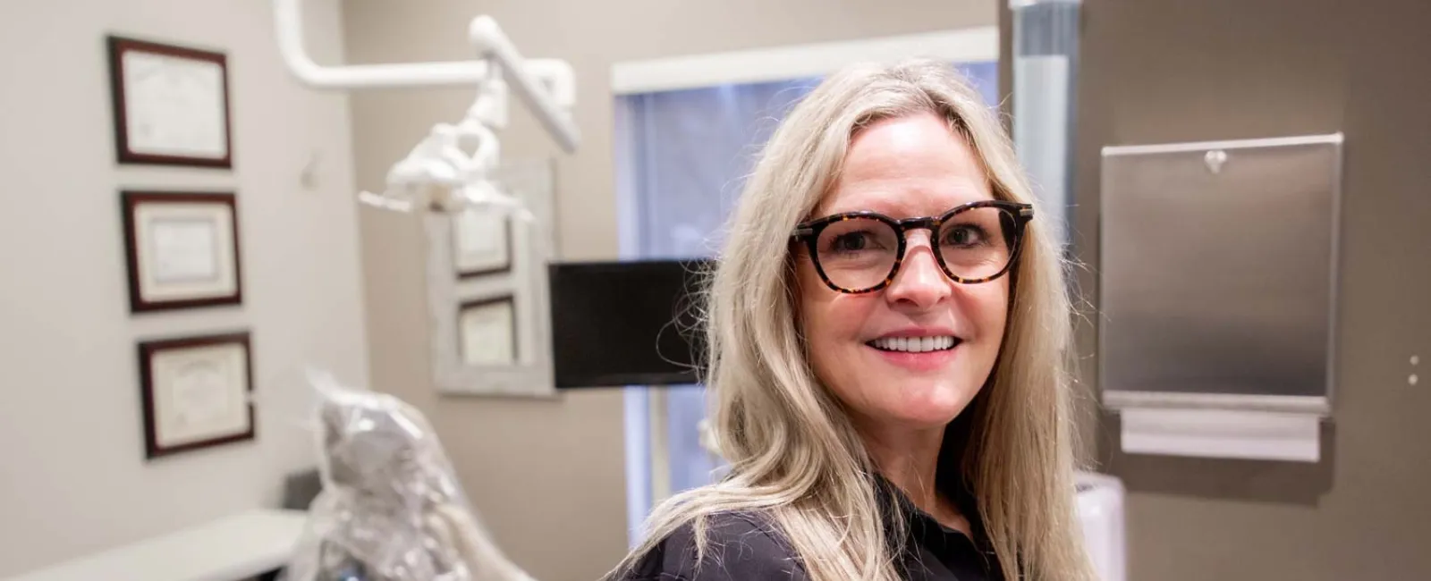 Smiling female dentist in black coat standing in a modern dental clinic with equipment and certificates.