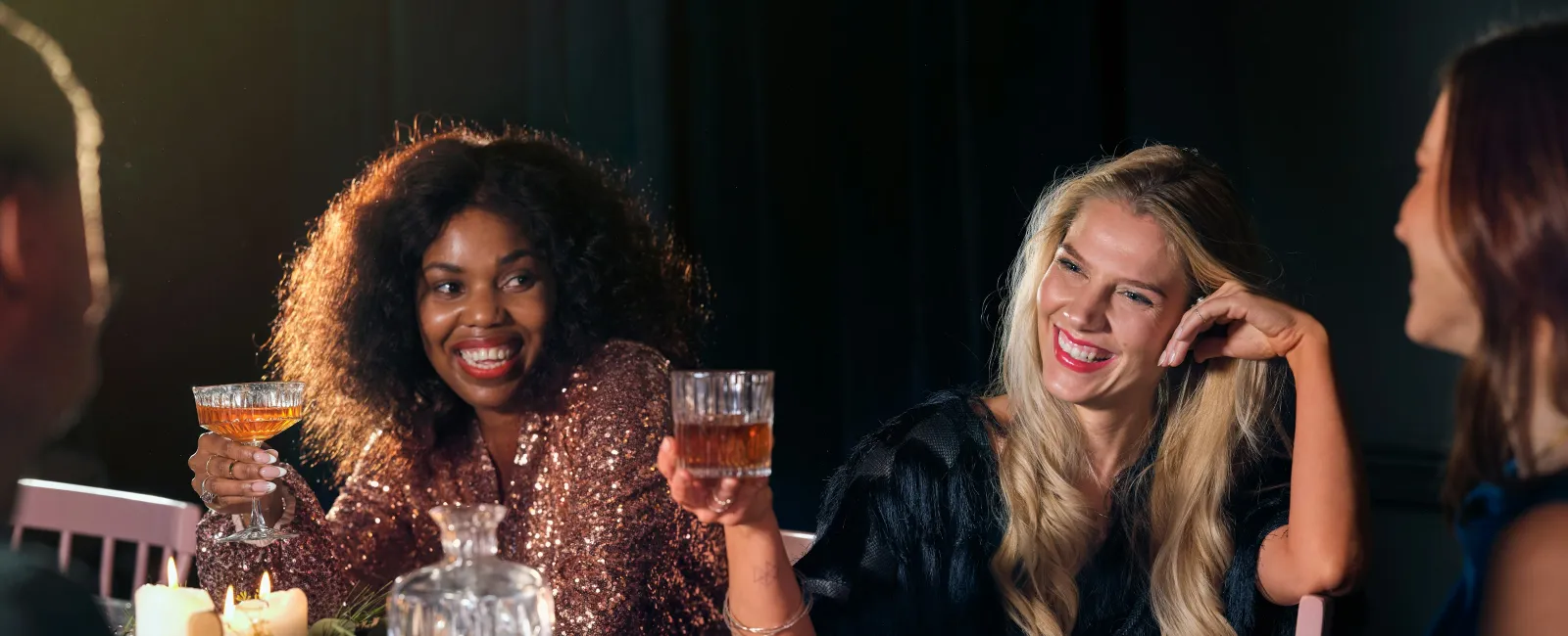 Group of friends smiling and toasting with drinks at a festive dinner table with candles and food.