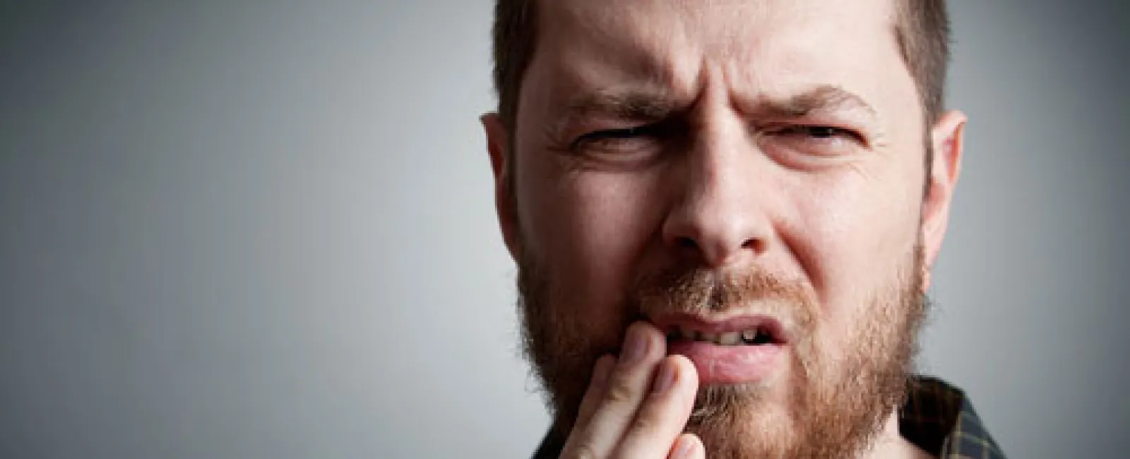Man with beard grimacing and holding his jaw in pain against a gray background