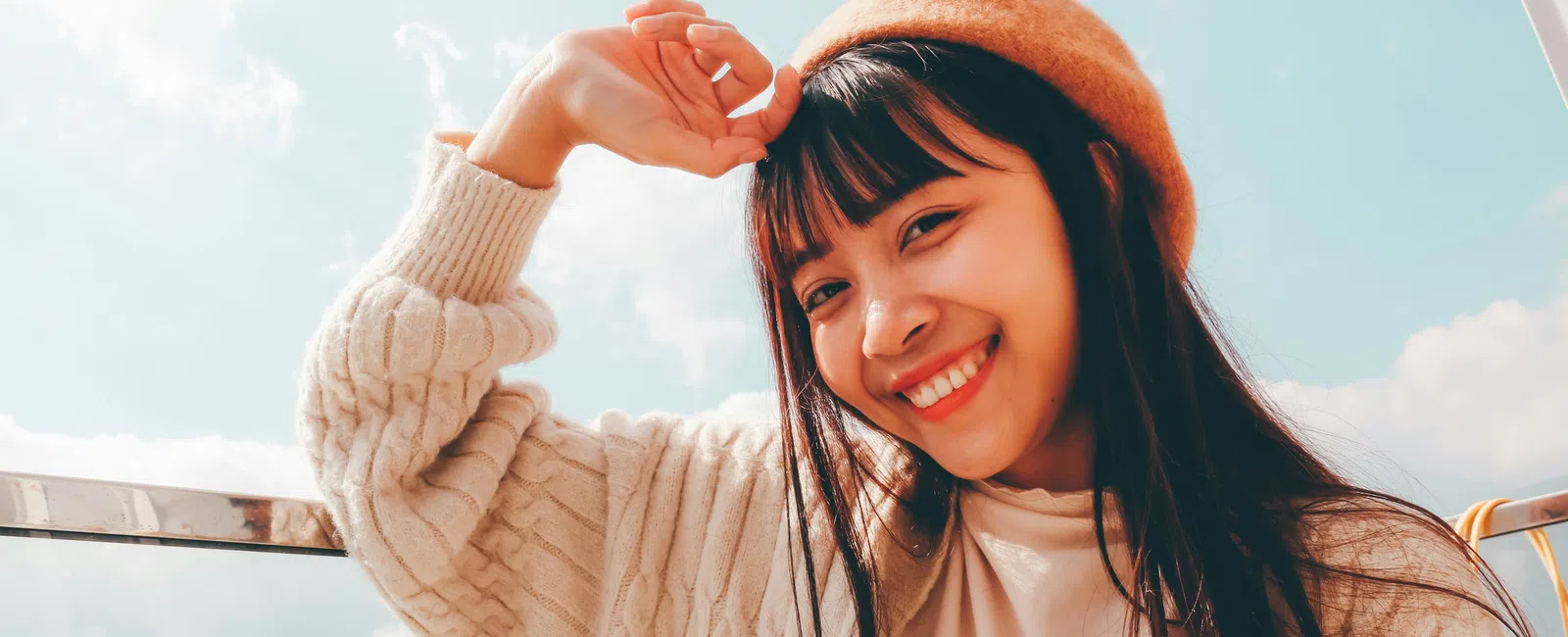 Young woman wearing an orange beret and sweater smiles outdoors under a blue sky with clouds