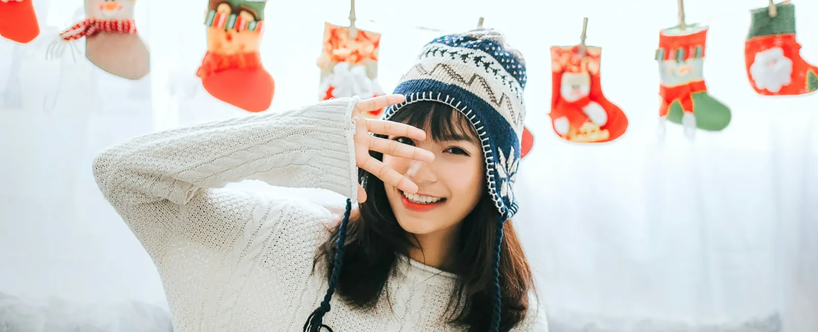 Young woman in cozy winter sweater and hat sitting on sofa with hanging Christmas stockings in background.