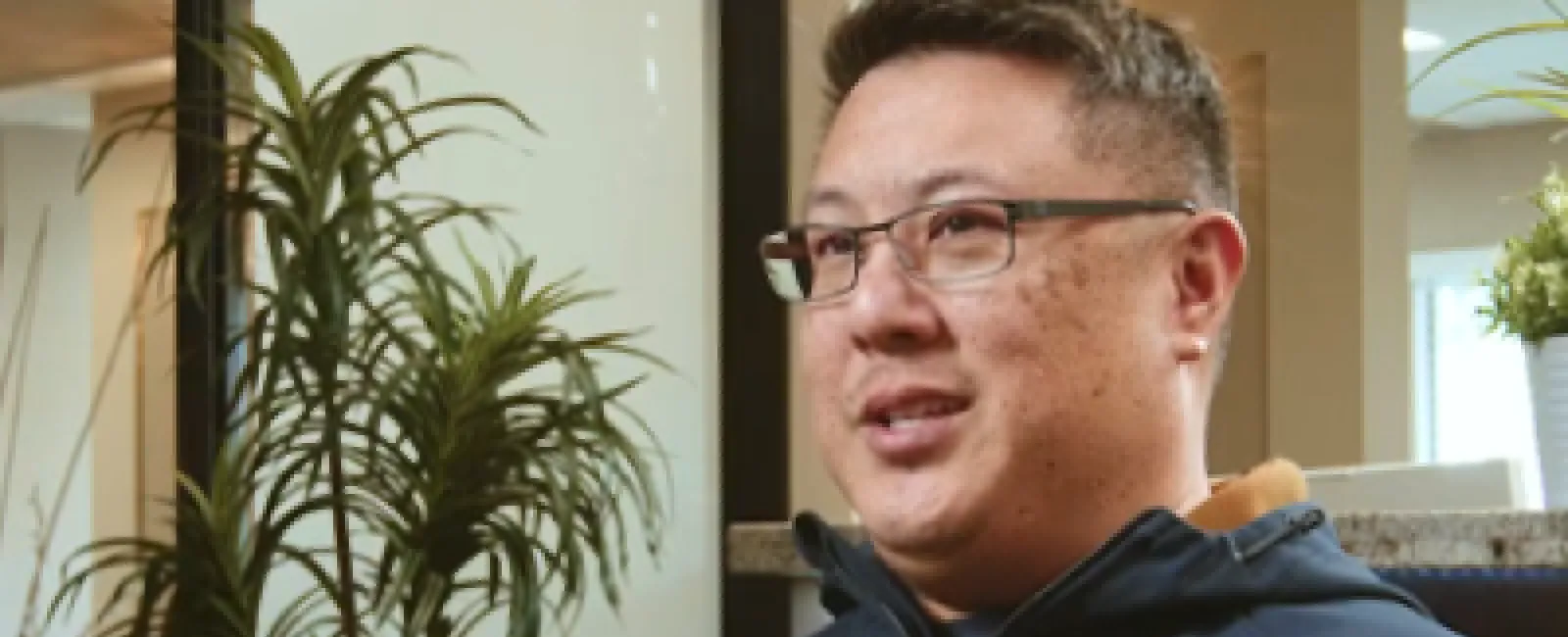Man with glasses wearing a navy blue zip-up jacket sitting indoors near a green potted plant.