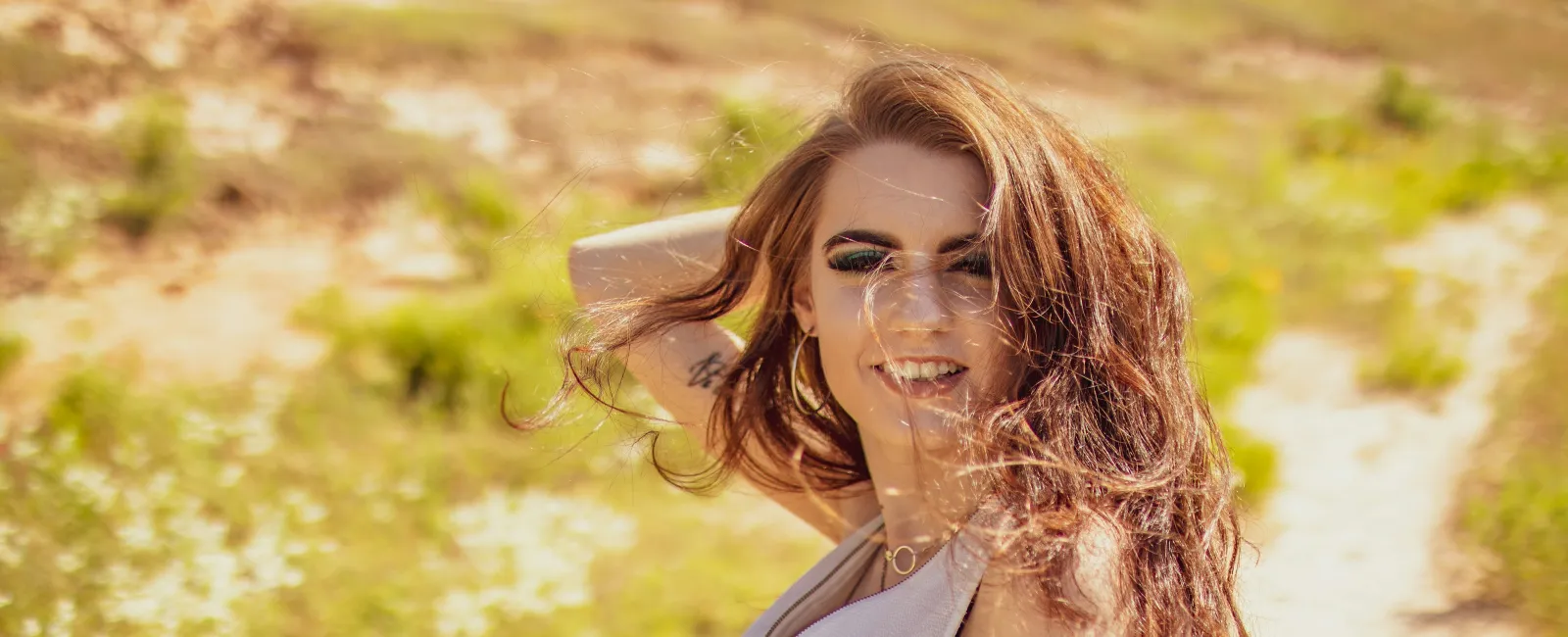 Smiling young woman with long flowing hair posing outdoors on a sunlit grassy path.