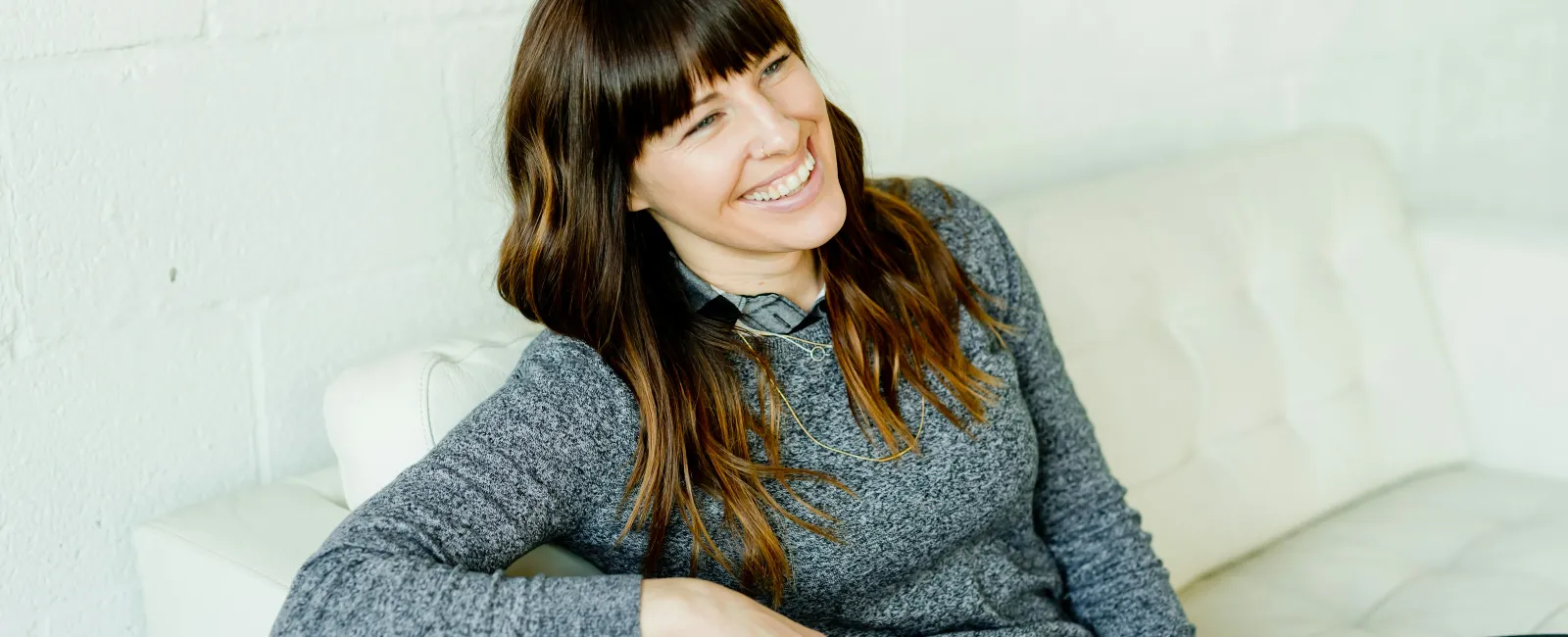 Smiling woman with long brown hair wearing a gray sweater sitting relaxed on a white couch indoors