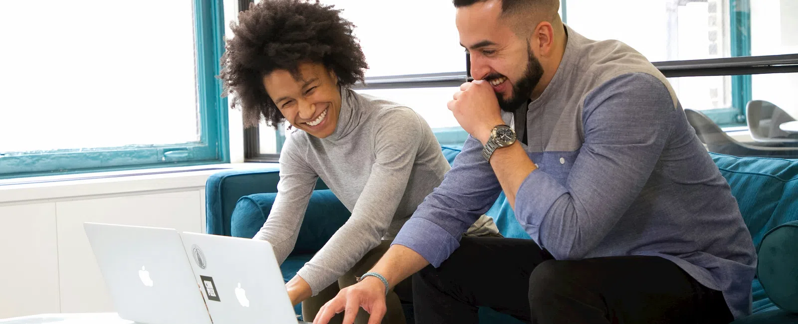 Two colleagues laugh while working together on laptops in a bright modern office lounge area.