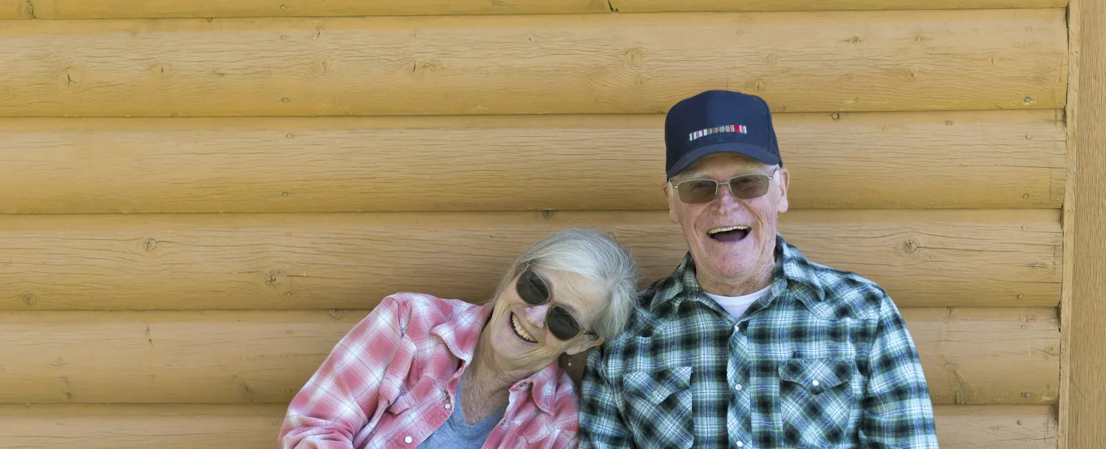 Elderly couple in plaid shirts sitting and smiling against a wooden log cabin wall on a sunny day