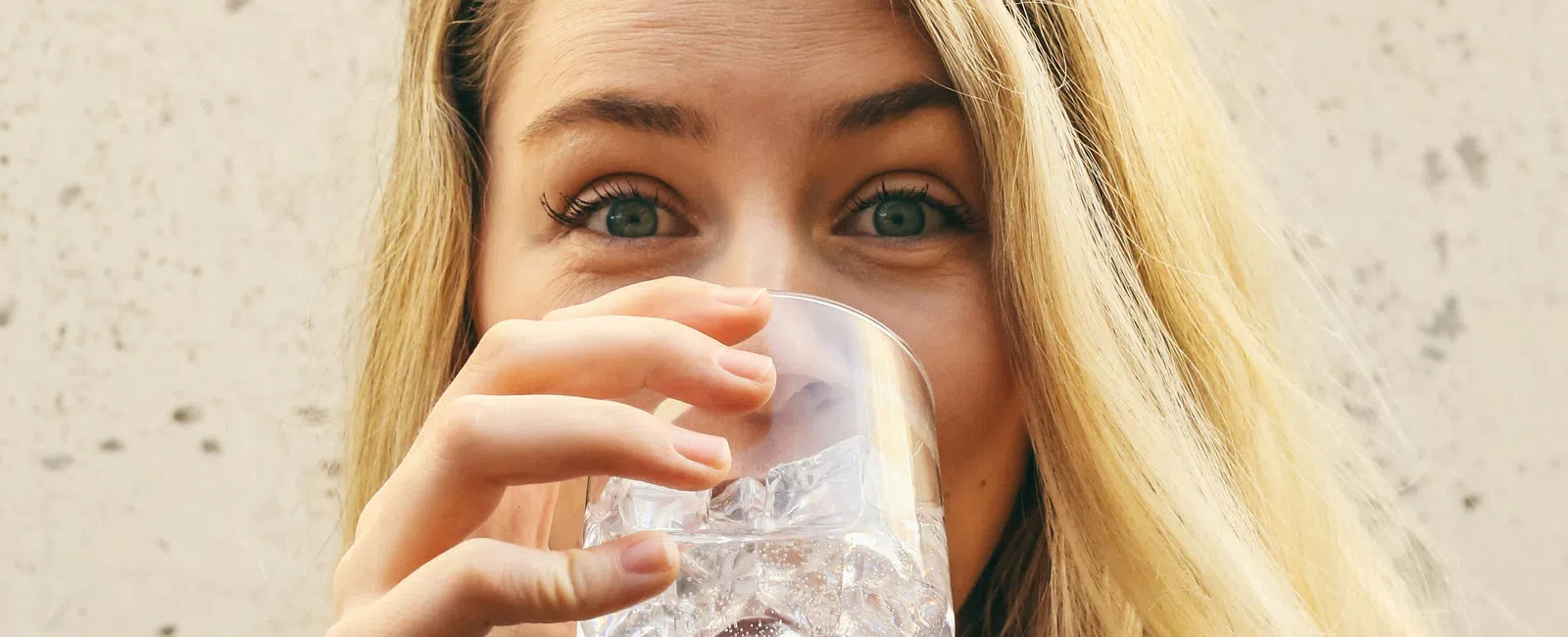 Young woman with long blonde hair drinking water from a clear glass filled with ice cubes.